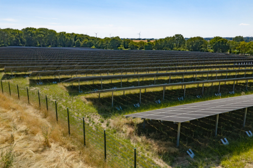 Luftaufnahme eines Solarparks im Gegenlicht mit Sicherheitszaun und Windrädern